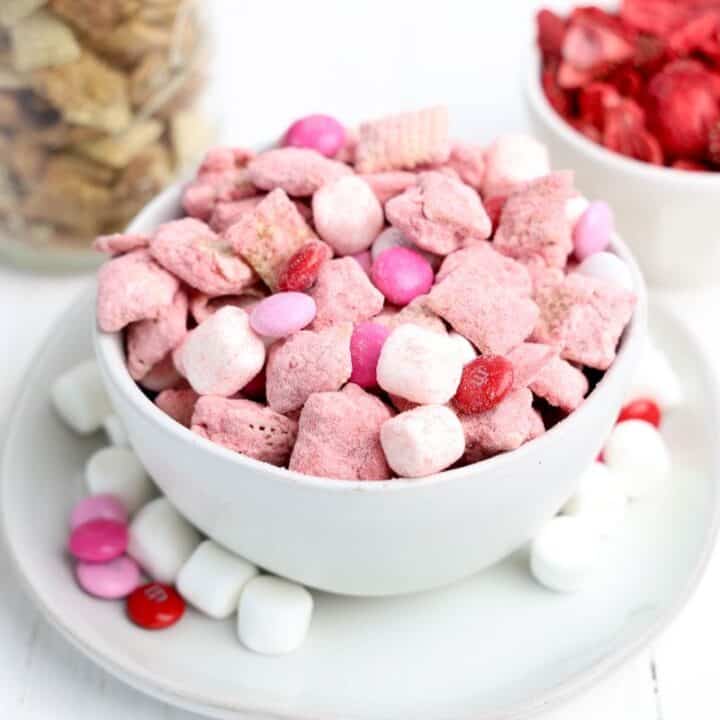 Small bowl of Valentine's Day Muddy Buddies with strawberry, white chocolate covered Chex cereal, M&Ms and mini marshmallows. With two small bowls of chex cereal and freeze dried strawberries in the background.