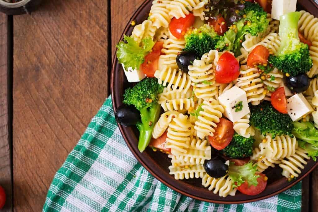Overhead view of a plate of summer pasta salad with broccoli, whole black olives, feta cubes and cherry tomatoes on a wooden table.