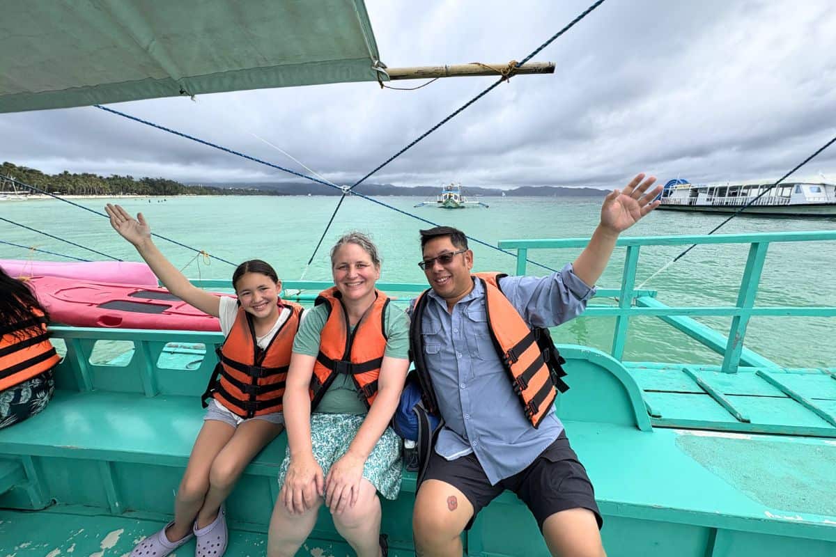 Three people wearing orange life jackets sit on a turquoise boat with their arms raised and smiling as they ride across calm turquoise water. A cloudy sky hangs over a tropical shoreline in the distance with other boats visible on the water, for a sunset cruise in Boracay. 