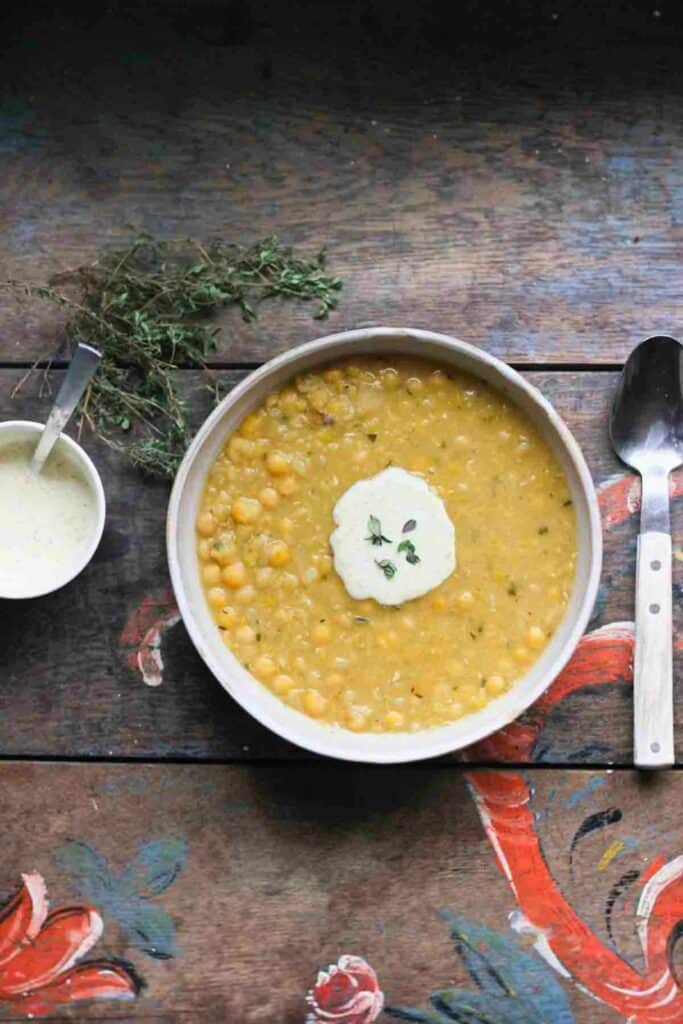 Vegetarian Swedish Ärtsoppa yellow pea soup is served in a white bowl on a rustic wooden table. The thick golden soup is topped with a dollop of cream and fresh herbs.