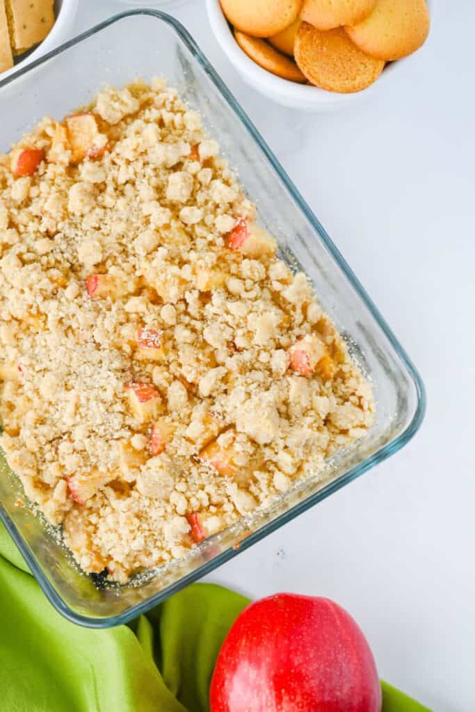 A glass baking dish is filled with a chunky apple crumble dessert dip, featuring tender apple pieces and a golden, buttery streusel topping. Surrounded by vanilla wafers and graham crackers, this warm and cozy Thanksgiving dip is ready for scooping and sharing.
