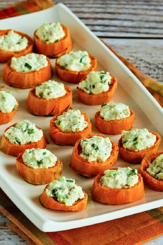 Roasted sweet potato rounds topped with a creamy herb and green onion mixture are arranged across a large white serving platter. The bite sized appetizers are served on an autumn colored cloth over a rustic wooden table.