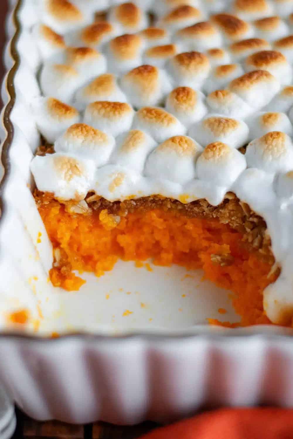 A close up of Sweet Potato Marshmallow Casserole with a creamy orange sweet potato layer, a crunchy brown sugar topping, and toasted mini marshmallows in a white baking dish with one serving scooped out.