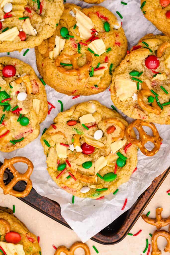 Close up of sweet and salty Christmas cookies on a parchment-lined baking sheet, packed with red and green M&Ms, white chocolate chunks, pretzels, potato chips, and festive holiday sprinkles.