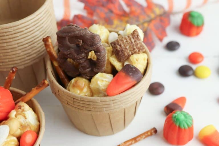 Small brown paper cup filled with a sweet and salty fall snack mix including caramel popcorn, chocolate-covered pretzel, candy corn, Chex cereal, bugels, pretzel sticks, and Reese’s Pieces, with candy pumpkins and fall leaves scattered around on a white surface.