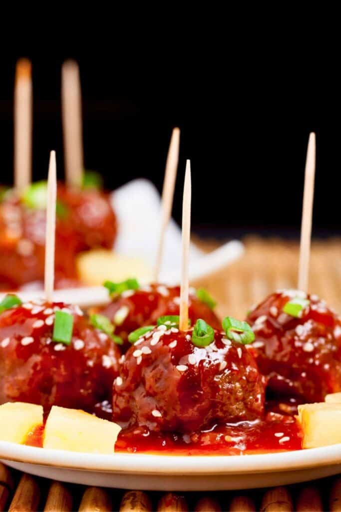Glazed sweet and sour meatballs served on a white plate, each topped with sesame seeds and chopped green onions, with toothpicks inserted for easy serving; chunks of pineapple add a fresh, tangy touch to the vibrant dish.