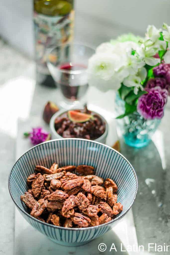 A bowl of spicy candied pecans in front of a wine bottle, glass of wine and pretty flowers.