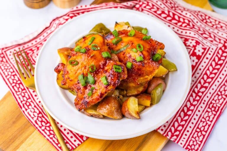 Golden-brown baked chicken thighs topped with chopped green onions, served over roasted red potatoes, green bell peppers, and onions on a white plate. The colorful chicken and veggies sheet pan dinner is set on a red and white patterned napkin with a gold fork beside it.