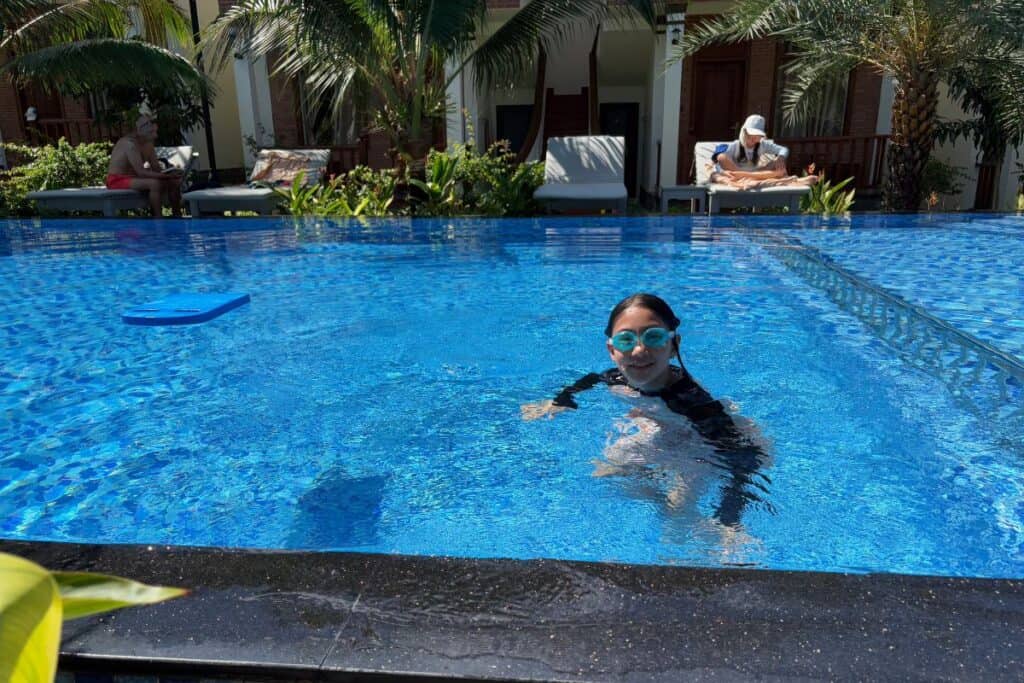 Child enjoying a swim in the clear blue pool at Palm Bay Resort, a family friendly resort in Phu Quoc, Vietnam. The background shows tropical palm trees, poolside loungers, and other guests relaxing under the sun near villa-style rooms.