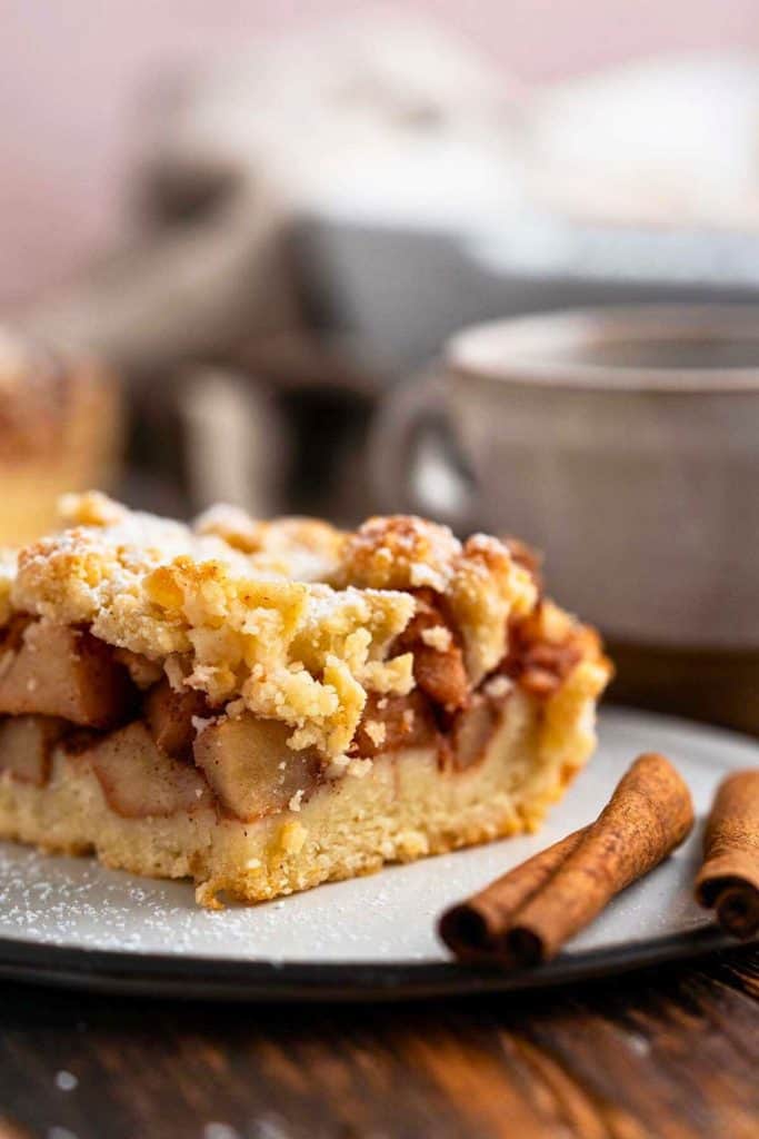 A close-up of a slice of apple crumb bar with a buttery crust, spiced apple filling, and golden crumble topping, lightly dusted with powdered sugar. It’s served on a plate next to cinnamon sticks and a warm mug, creating a cozy, autumn-inspired scene.