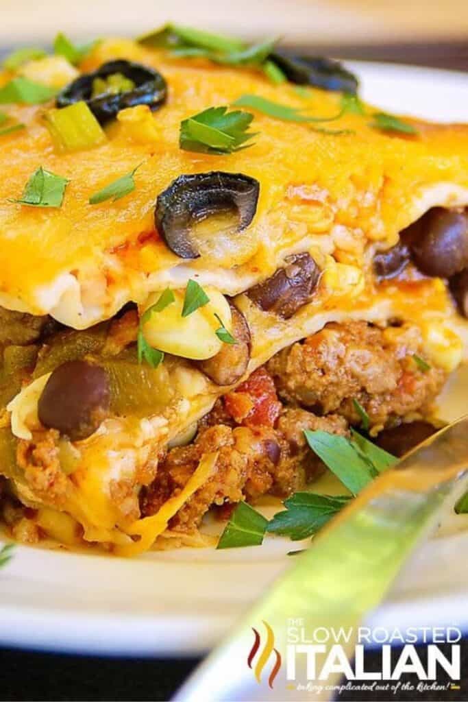 Slice of taco lasagna made with tortillas layered with seasoned ground beef, beans, vegetables, and melted cheese topped with black olives and cilantro.