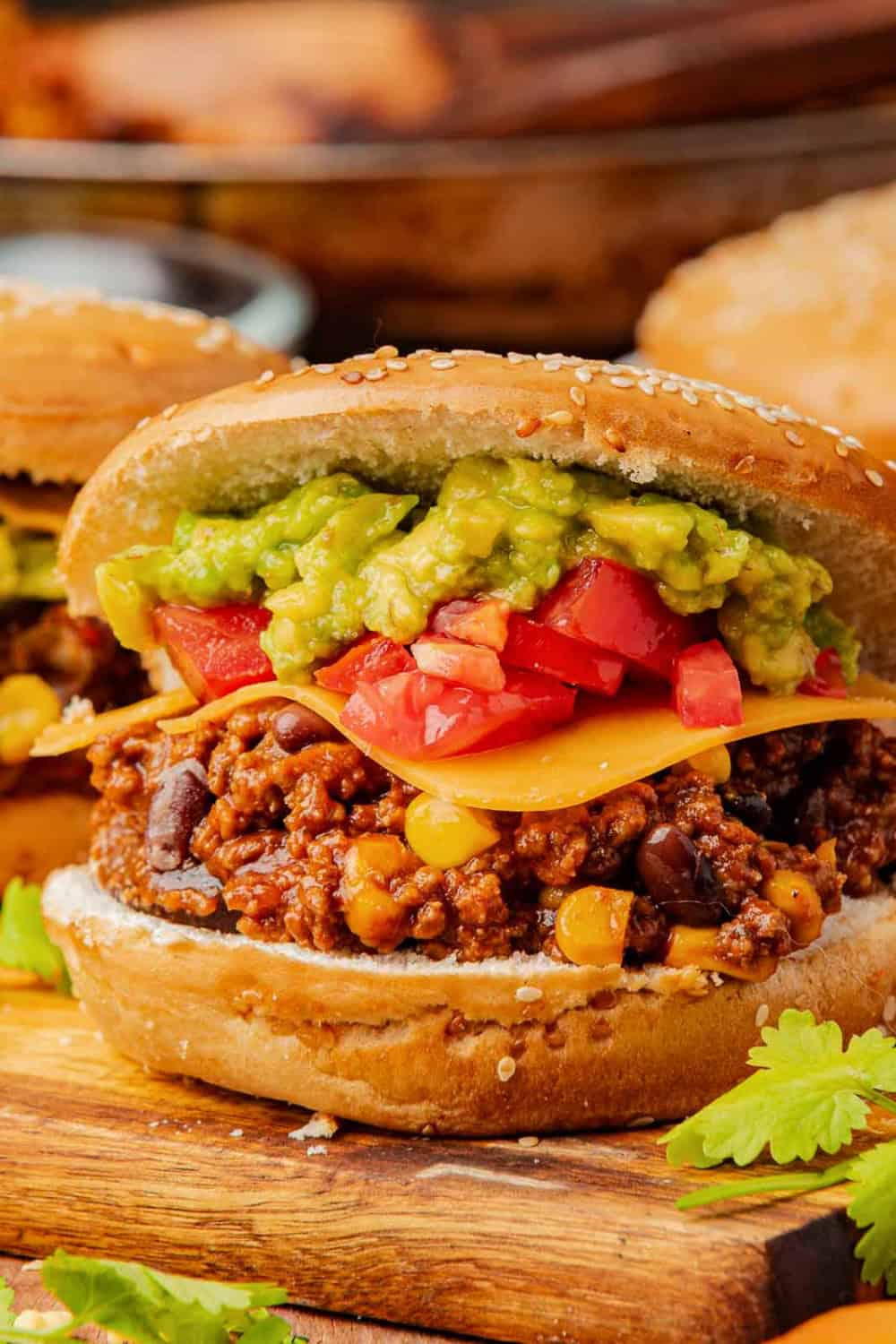 Taco Sloppy Joe on a sesame seed bun topped with seasoned ground beef, black beans, corn, cheddar cheese, diced tomatoes, and chunky guacamole. The sandwich is served on a wooden board with fresh cilantro scattered around it.