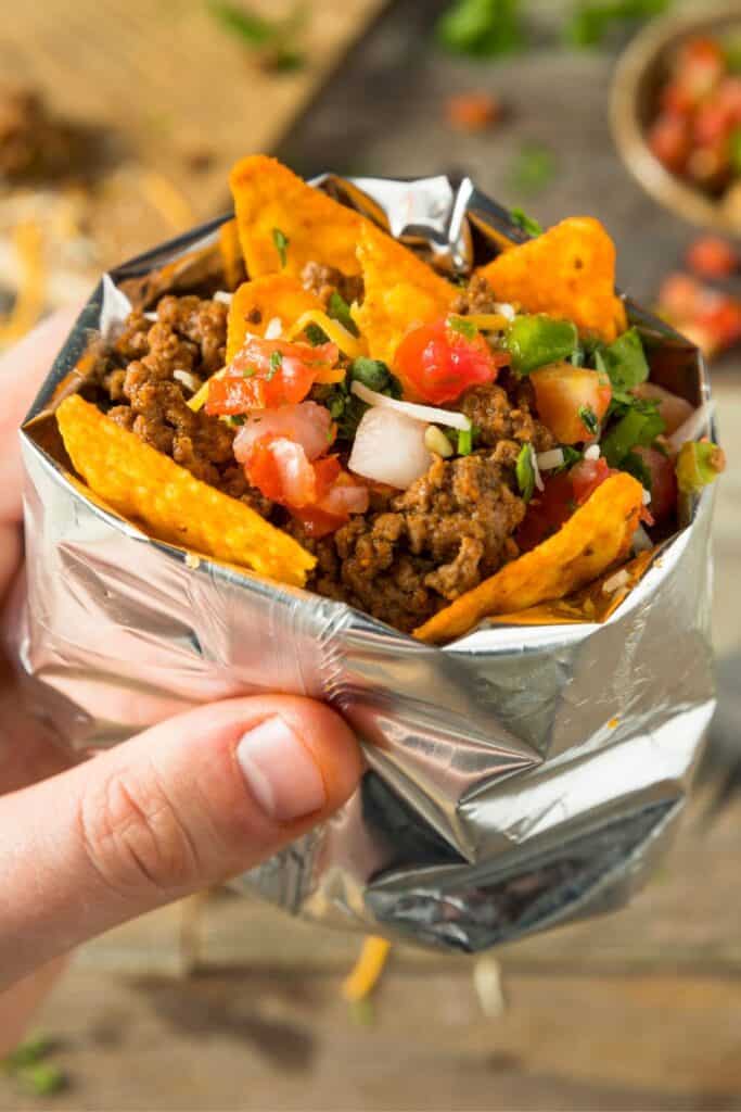 Hand holding a taco in a bag with dorito chips, ground beef, tomatoes and onions.