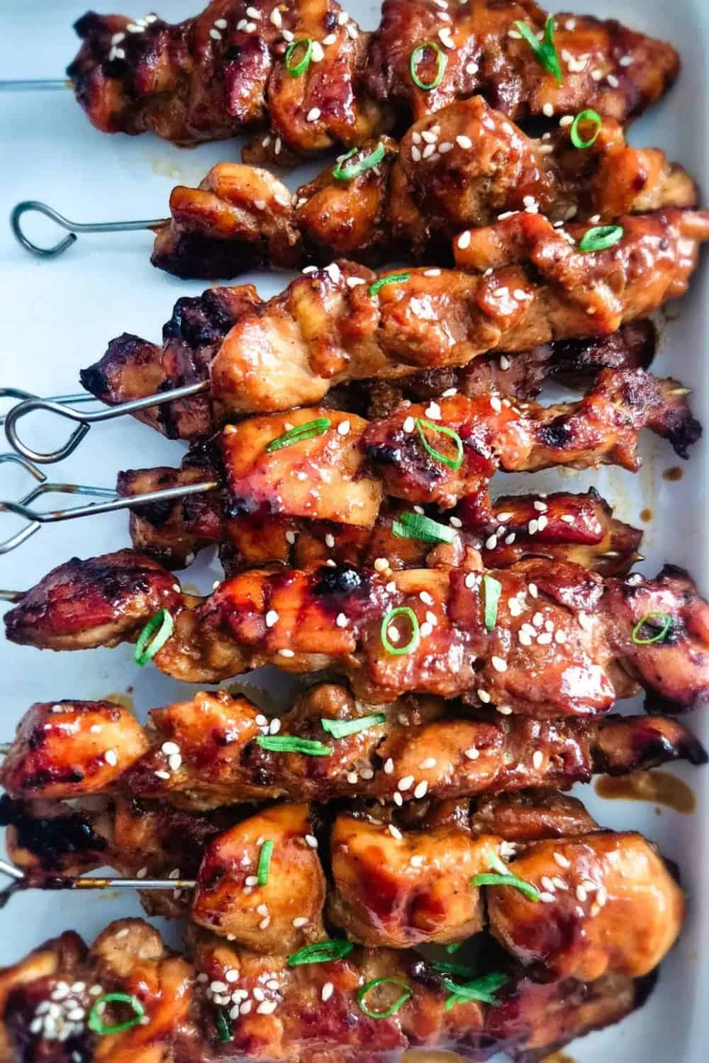 Teriyaki chicken skewers coated in a glossy sauce with charred edges. The grilled chicken is topped with white sesame seeds and sliced green onions.