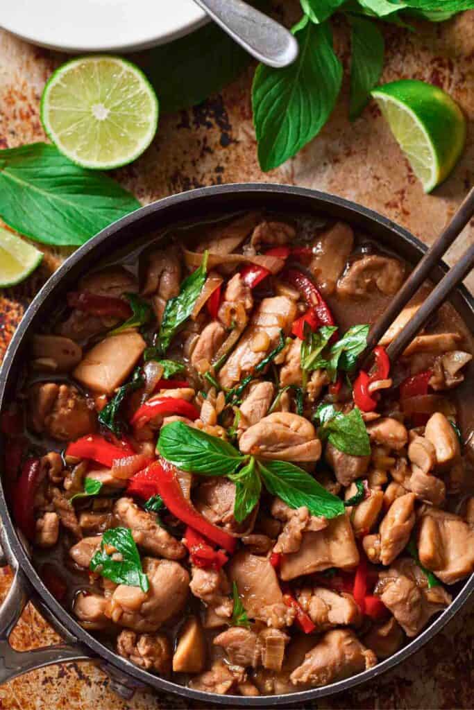 Thai basil chicken stir fry with tender chicken pieces cooked in a savory sauce with red peppers, onions, and fresh basil leaves. The dish is served in a pan with lime wedges and basil nearby highlighting the fragrant Thai basil chicken.