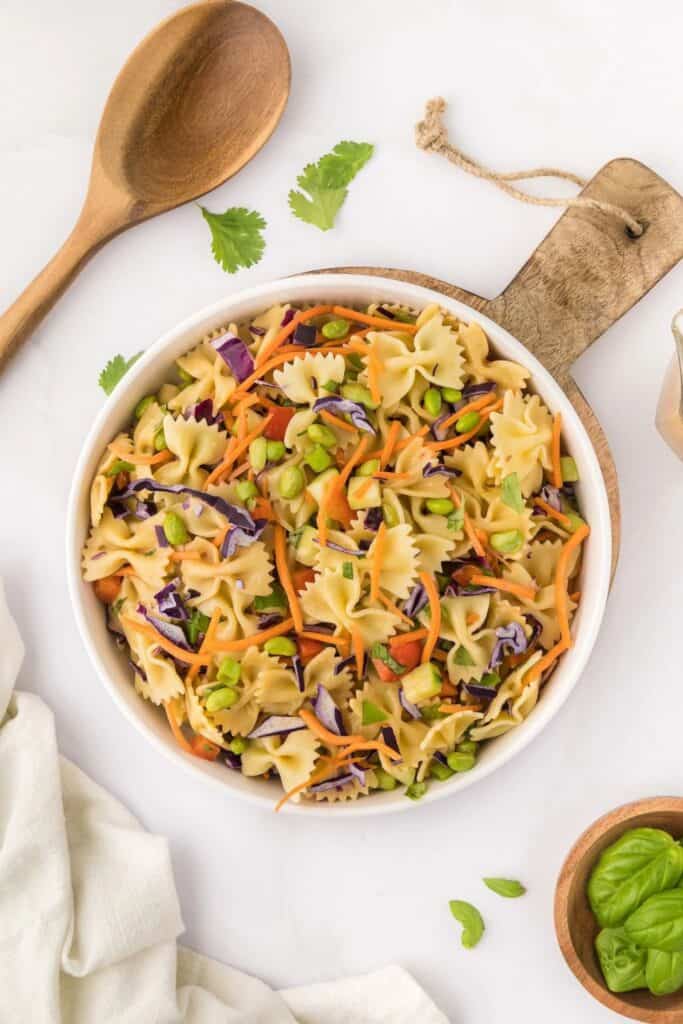 An overhead view of a bowl of bow tie Thai pasta salad with shredded carrots & cabbage, edamame, diced cucumbers, red peppers. 