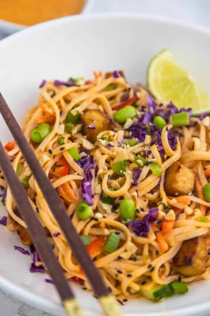 Bowl of Thai noodle salad with spaghetti noodles edamame shredded carrots purple cabbage green onions and crispy tofu tossed in spicy peanut sauce and topped with sesame seeds and chopped peanuts. Lime wedge and chopsticks beside the bowl suggest a fresh Asian inspired noodle salad
