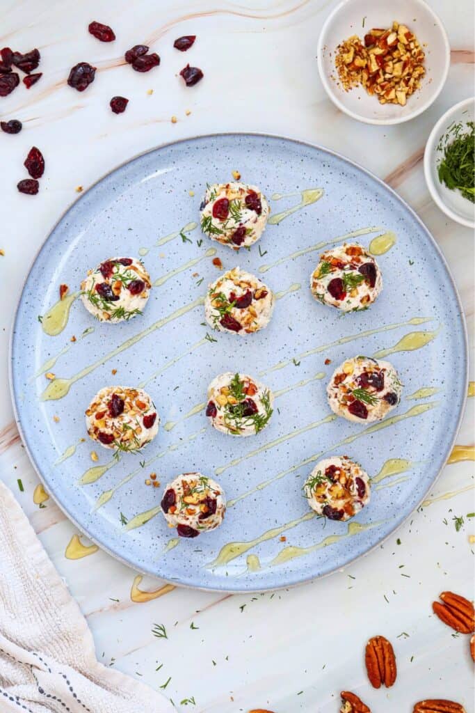 Eight cranberry pecan cheese balls topped with dried cranberries. Chopped pecans and fresh dill are spaced across a large pale blue platter with honey drizzled between them. Bowls of extra pecans and dill and scattered cranberries and whole pecans surround the easy cold appetizer.