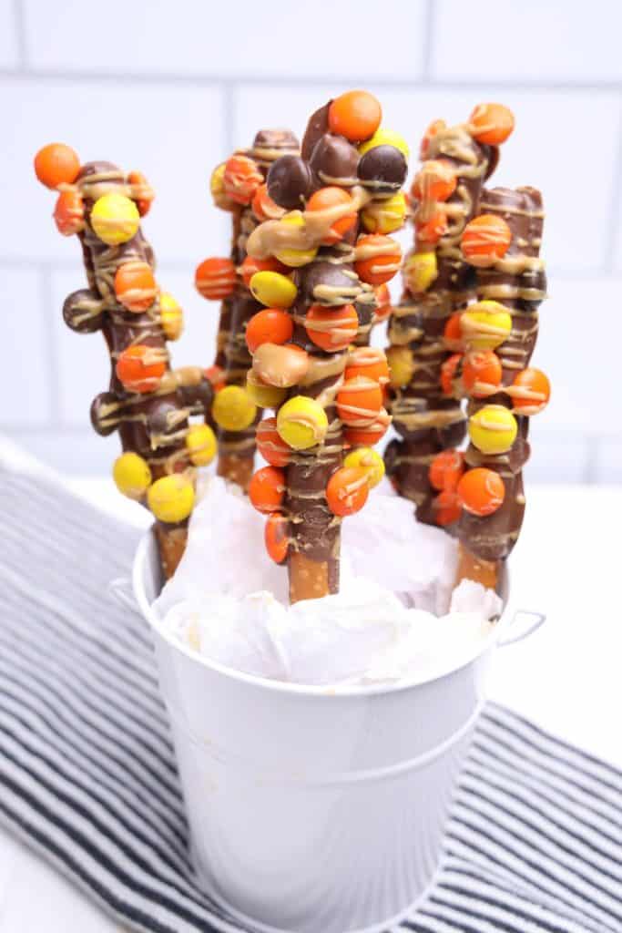 White pail filled with Thanksgiving Chocolate Covered Pretzels with Reese's Pieces, featuring pretzel rods dipped in chocolate, coated in orange, yellow, and brown candy, and drizzled with peanut butter.