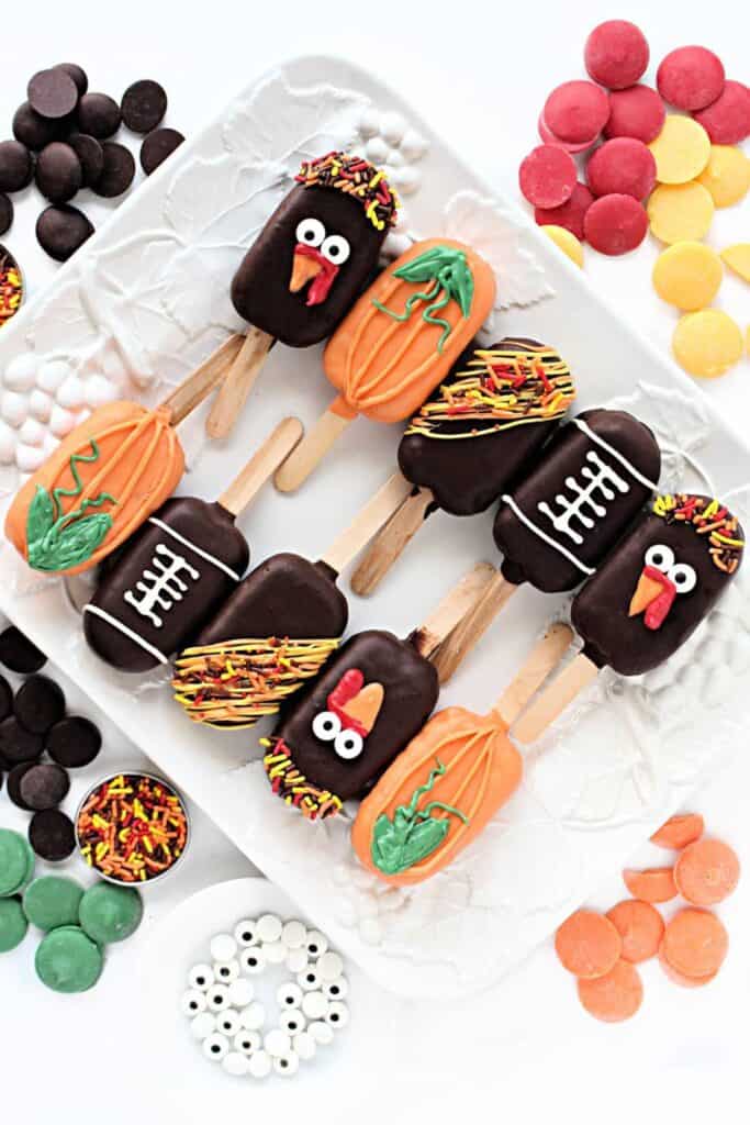 Thanksgiving Peanut Chews Candy Pops decorated as orange pumpkins, chocolate turkeys, footballs, and festive sprinkle covered treats. The candy pops are arranged on a white platter surrounded by colorful candy melts, sprinkles, and candy eyes.