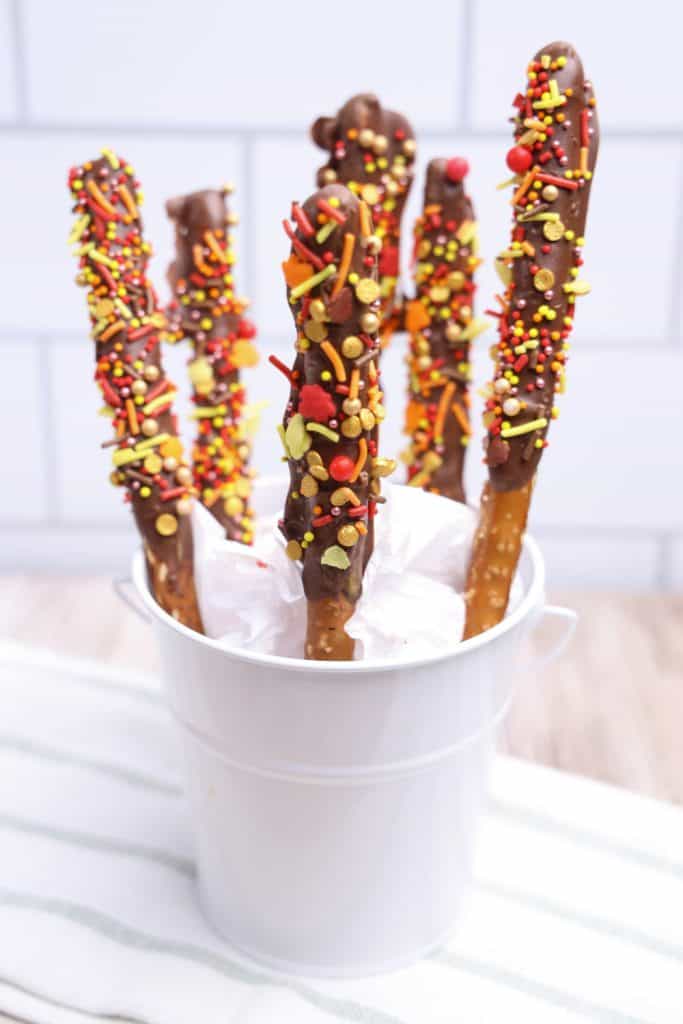 White pail filled with Thanksgiving pretzel rods coated in chocolate and topped with festive fall-colored sprinkles in red, yellow, and orange. The rods are propped up in white tissue paper, highlighting their colorful autumn-themed decoration.