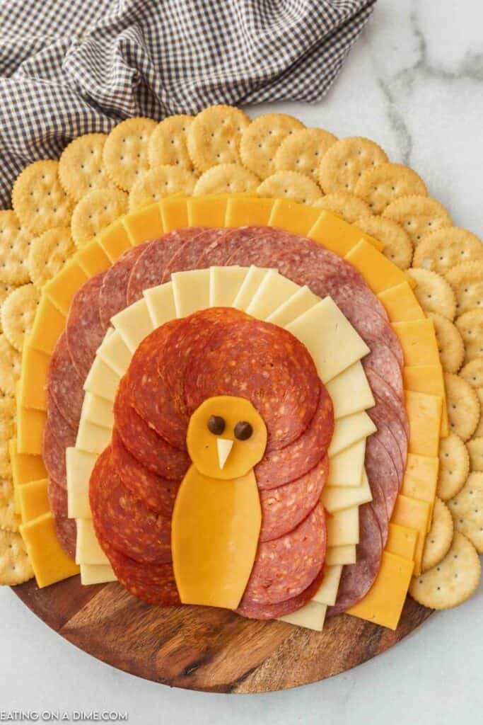 Thanksgiving turkey charcuterie board features overlapping layers of crackers, cheddar cheese, salami, white cheese, and pepperoni arranged like colorful tail feathers. Slices of cheddar form the turkey&rsquo;s body and head, with small dark eyes and a white cheese beak completing the festive design.