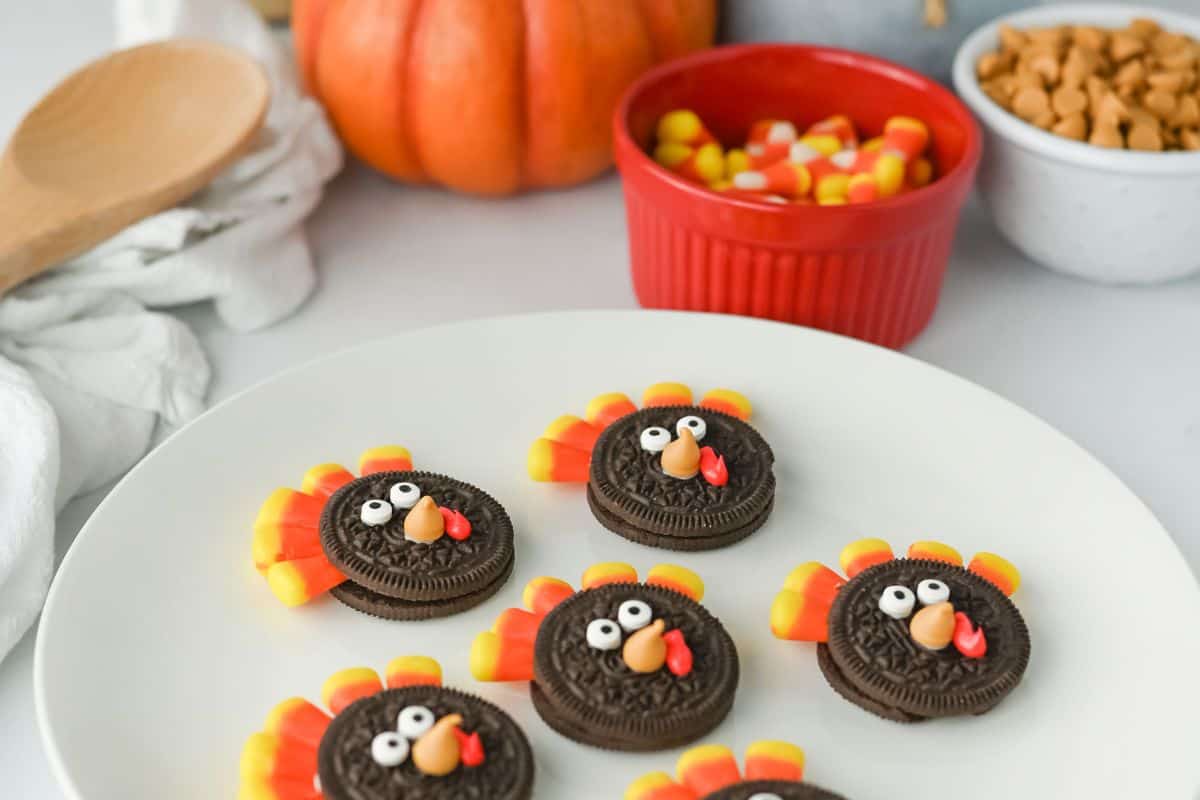 Plate of festive Turkey Oreos made from chocolate sandwich cookies, candy eyes, candy corn feathers, and a small orange candy beak with red icing wattle. A pumpkin and bowls of candy corn and butterscotch chips are visible in the background, adding to the Thanksgiving theme.