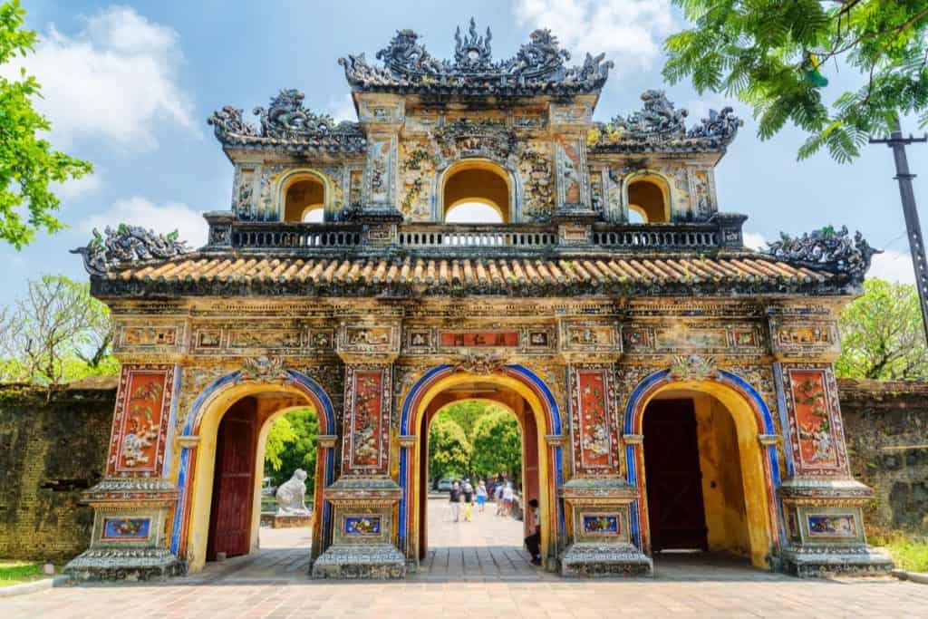 A richly decorated gate with three arched entrances stands at the Imperial City in Hue, Vietnam, featuring intricate carvings, colorful mosaics, and dragon motifs. The weathered structure is framed by greenery under a bright blue sky, with visitors walking through the central archway.