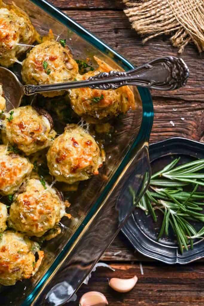 Golden baked stuffed mushrooms topped with melted cheese and herbs sit in a glass baking dish with a serving spoon resting across the top. Fresh rosemary and garlic are arranged beside the dish on a rustic wooden table for a warm spread of Thanksgiving Day appetizers.