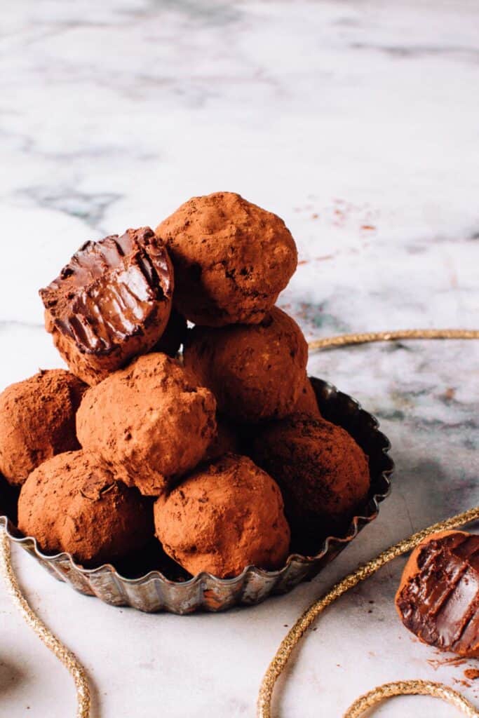 Rich chocolate truffles coated in cocoa powder, stacked in a fluted metal dish with one truffle revealing its smooth, glossy ganache center.