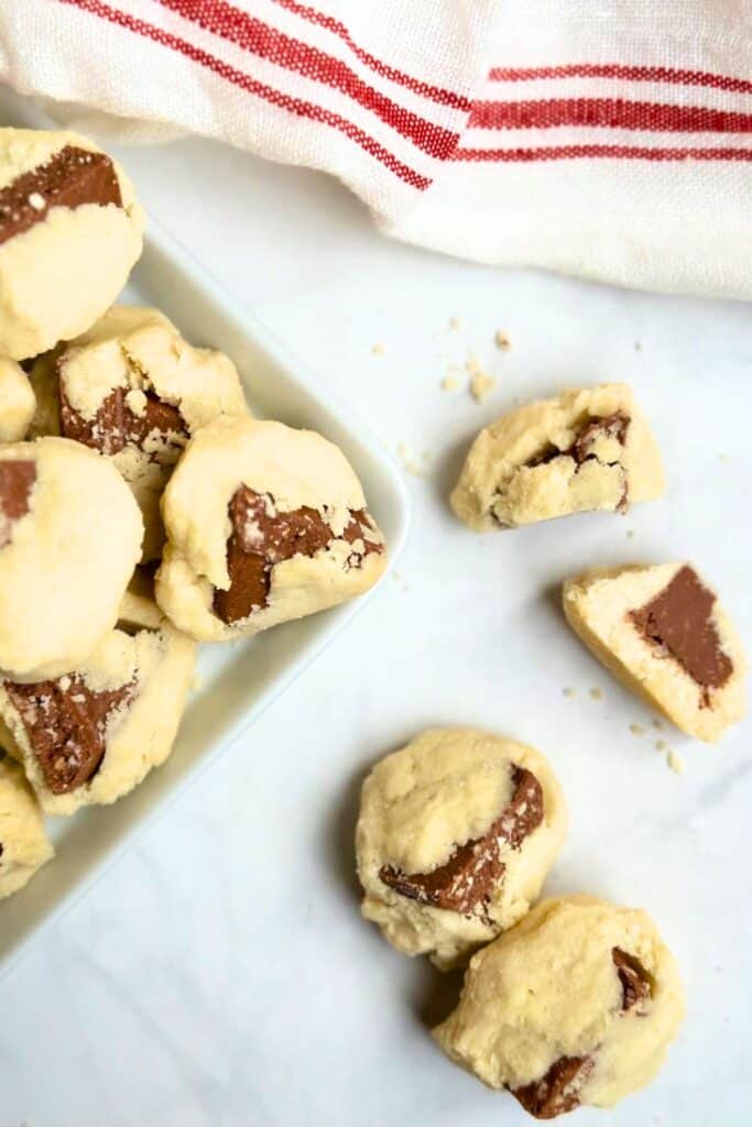 Soft, buttery shortbread cookies with chunks of Toblerone chocolate peeking through the dough. The cookies are displayed on a white surface next to a red-striped kitchen towel, with a few broken pieces showing the creamy chocolate center.