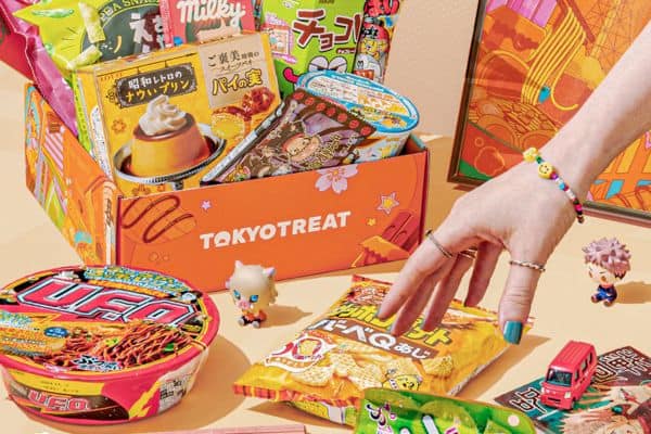Brightly colored TokyoTreat subscription box filled with assorted Japanese snacks, including ramen bowls, chips, candies, and chocolates, with anime-themed packaging. A hand with colorful rings and a beaded bracelet reaches for a snack, surrounded by miniature toy figures and a small red bus, creating a playful and vibrant unboxing scene.