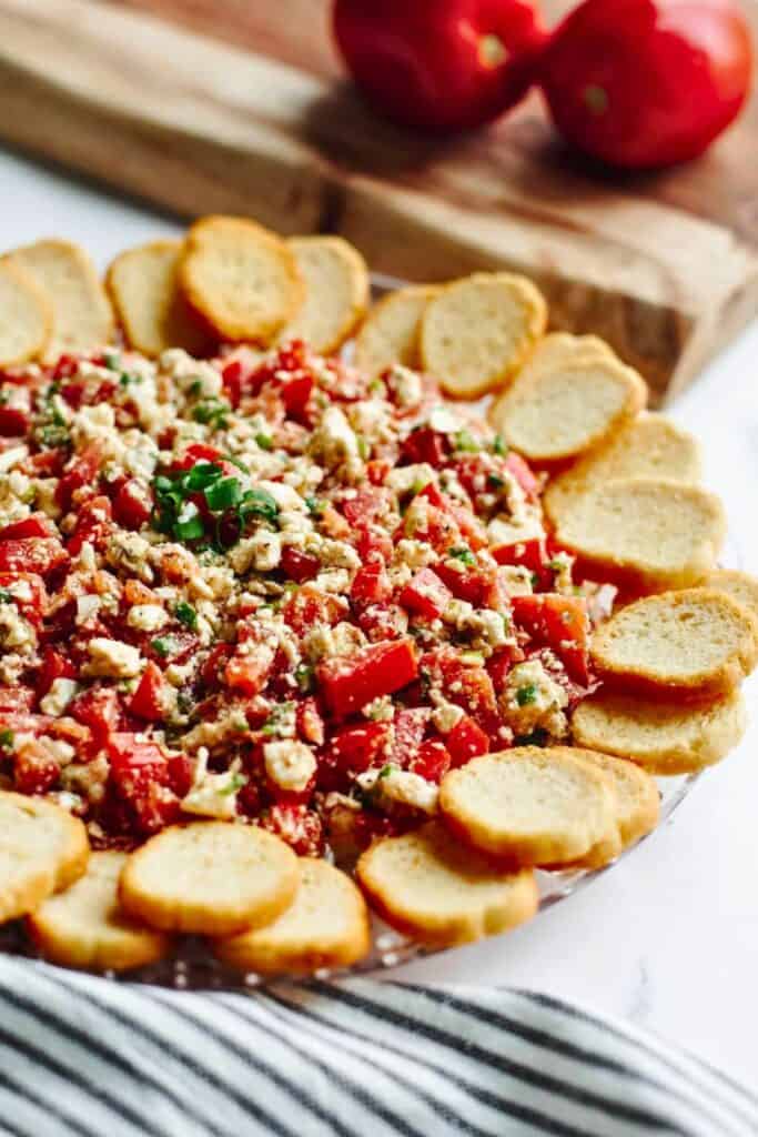 Tomato feta dip made with chopped fresh tomatoes, crumbled feta cheese, herbs, and green onions is piled in the center of a serving platter. Toasted baguette slices are arranged around the dip for scooping and serving.