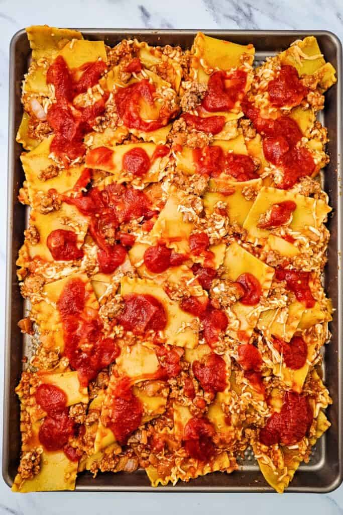 Sheet pan lasagna topped with spoonfuls of tomato pasta sauce over noodles mixed with Italian sausage and cheese. The extra sauce is scattered across the pan before the final toppings are added.