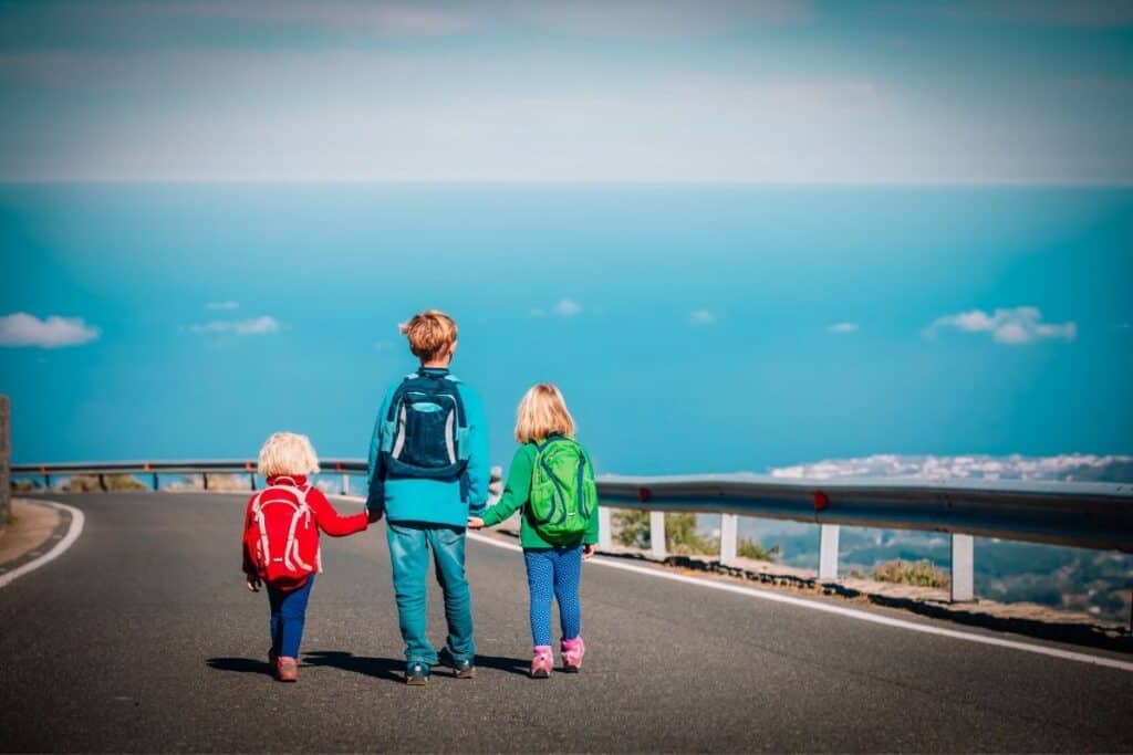 3 children walking hand in hand down road wearing travel backpacks