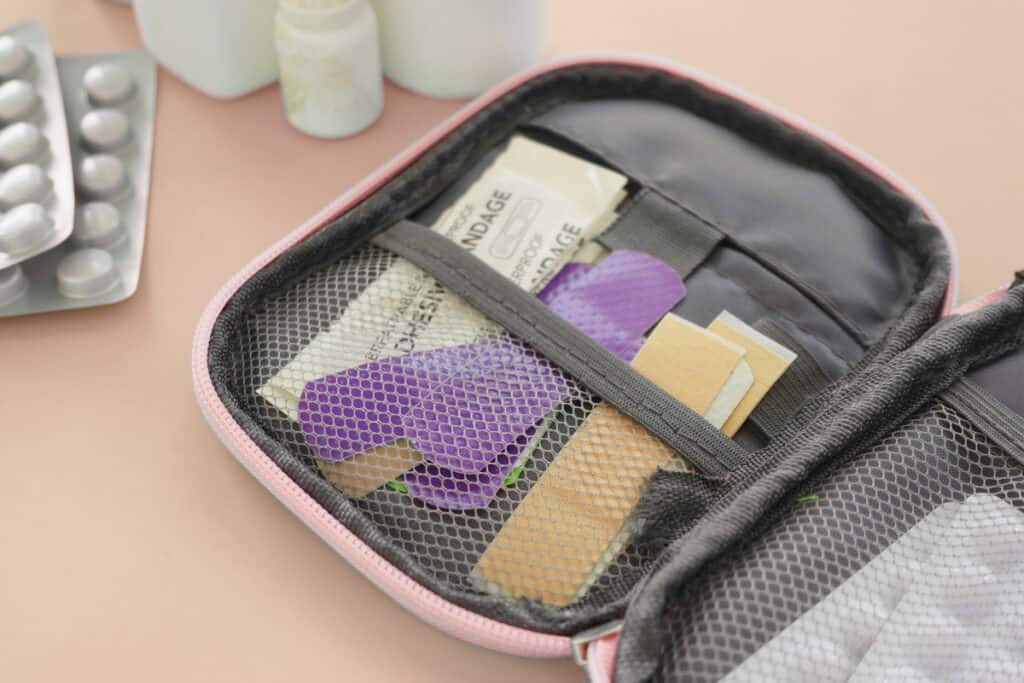Open travel first aid kit with mesh compartments holding adhesive bandages blister pads and wrapped gauze alongside pill bottles in the background.