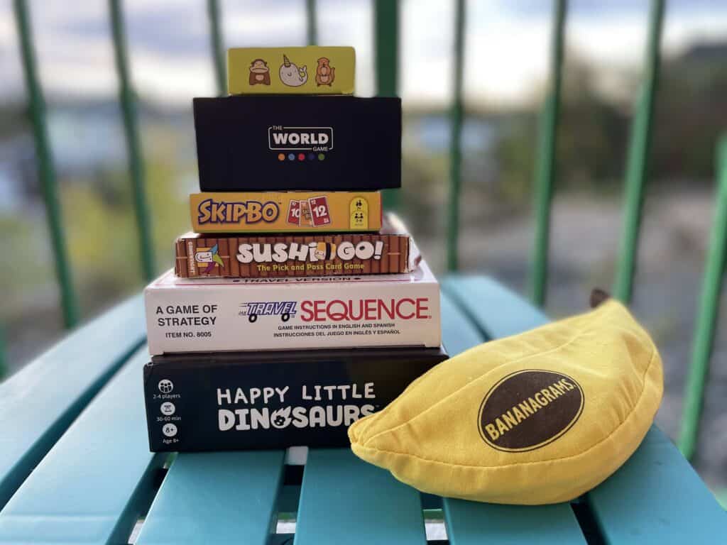 7 Travel game ideas for tweens, on a small outdoor side table. Happy Little Dinosaur, Bananagrams, Travel sequence, Sushi Go, Skip-Bo, The world Game.  
