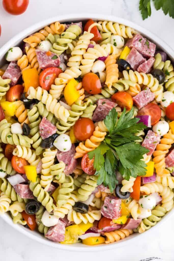 Colorful bowl of tri-color rotini pasta salad mixed with cherry tomatoes, yellow bell peppers, salami chunks, black olives, red onion, mozzarella balls, and fresh parsley. The ingredients are tossed with a light Italian dressing and served in a white bowl.