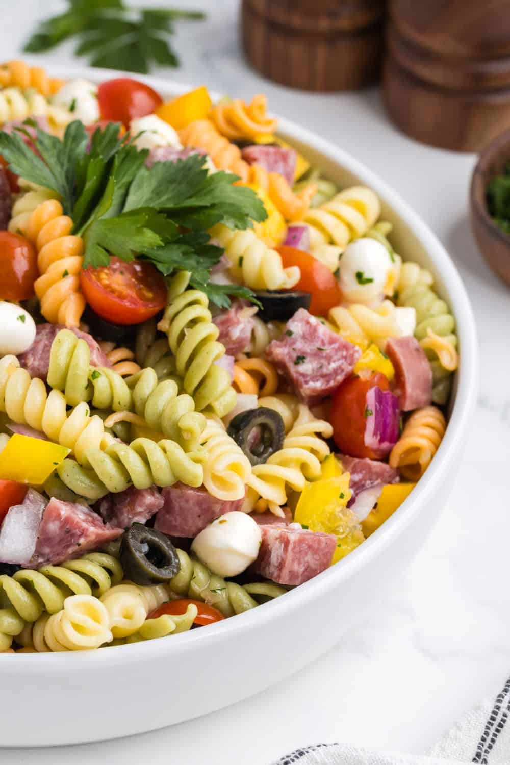 Colorful tri-color rotini pasta salad served in a white bowl, loaded with cherry tomatoes, cubed salami, mozzarella balls, black olives, red onion, and yellow bell pepper, all tossed in Italian dressing and topped with fresh parsley, perfect as a cookout side dish.