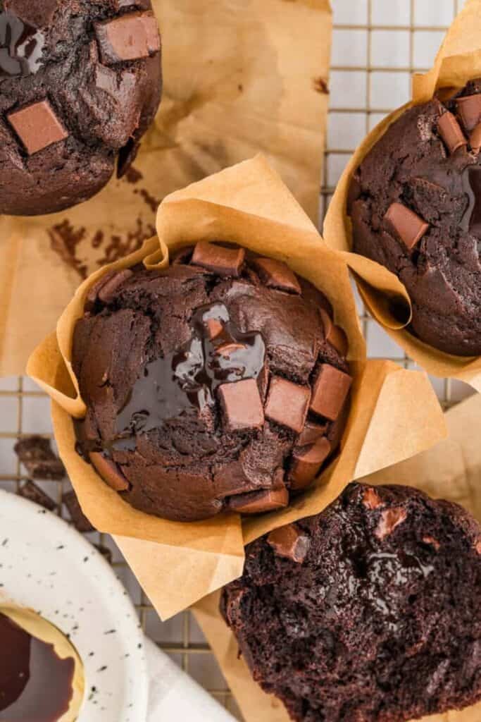Overhead view of rich triple chocolate muffins wrapped in brown parchment liners, topped with chocolate chunks and glossy pools of melted chocolate. One muffin is sliced open to reveal its dense, moist interior packed with gooey chocolate.