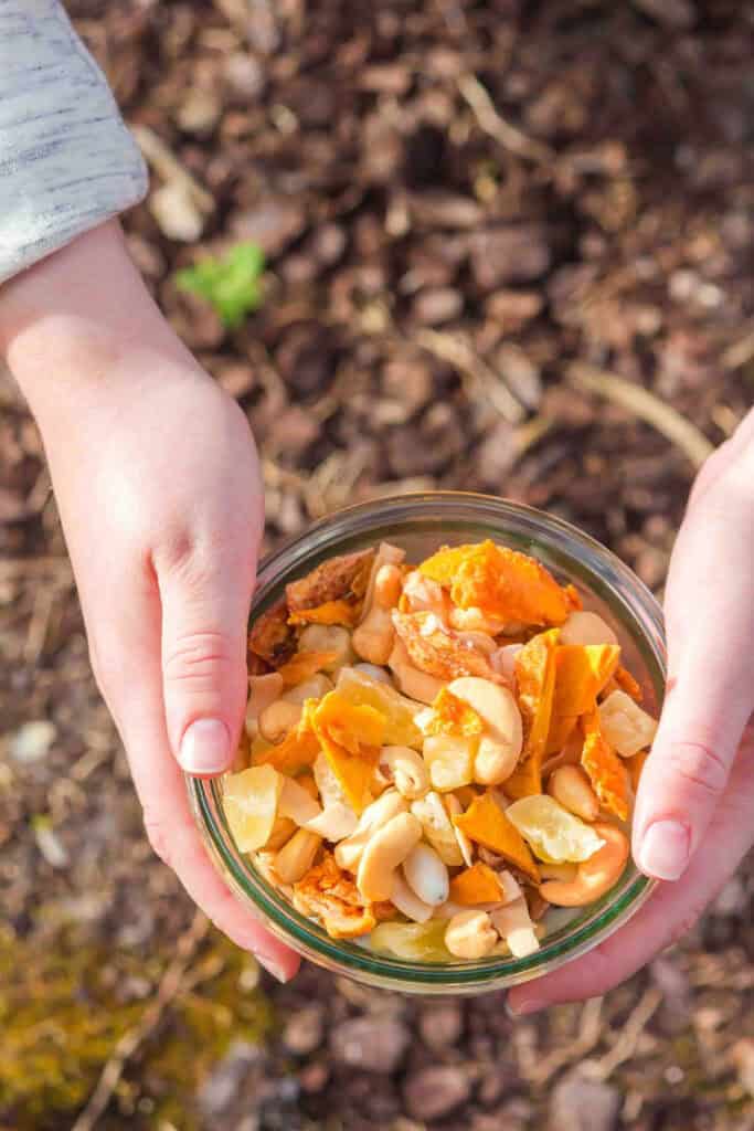 Hands holding a glass bowl of tropical trail mix, outdoors over a path with leaves. 