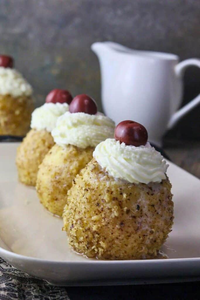 Three Bosnian-style poached apples are plated in a row, each coated in finely ground walnuts and topped with a swirl of whipped cream and a red grape. This elegant fall dessert offers a unique and festive twist for Thanksgiving, blending tender fruit with rich, nutty flavors.