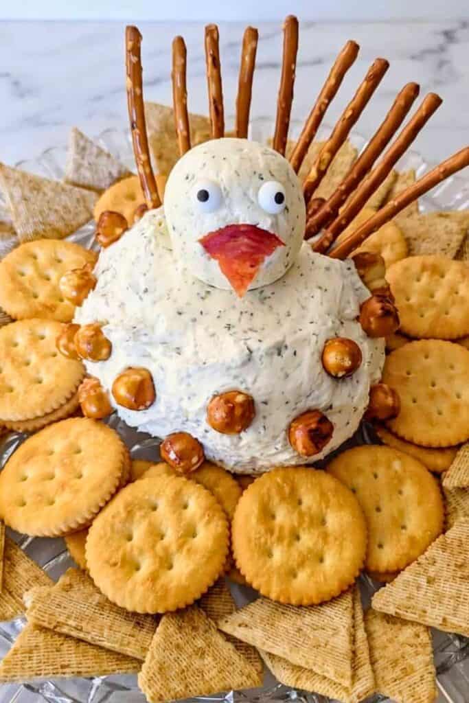 A turkey cheeseball decorated with candy eyes and a red and orange pepper beak sits in the center of a serving platter. Pretzel sticks fan out behind the cheeseball like turkey feathers, while crackers, pretzels, and crunchy snacks surround it for dipping.