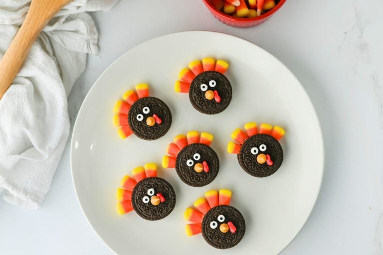 Overhead view of a white plate holding six Oreo Turkey Cookies decorated with candy eyes, candy corn feathers, orange candy beaks, and red icing wattles. A bowl of candy corn and a wooden spoon rest nearby, adding to the playful, fall-themed setting.