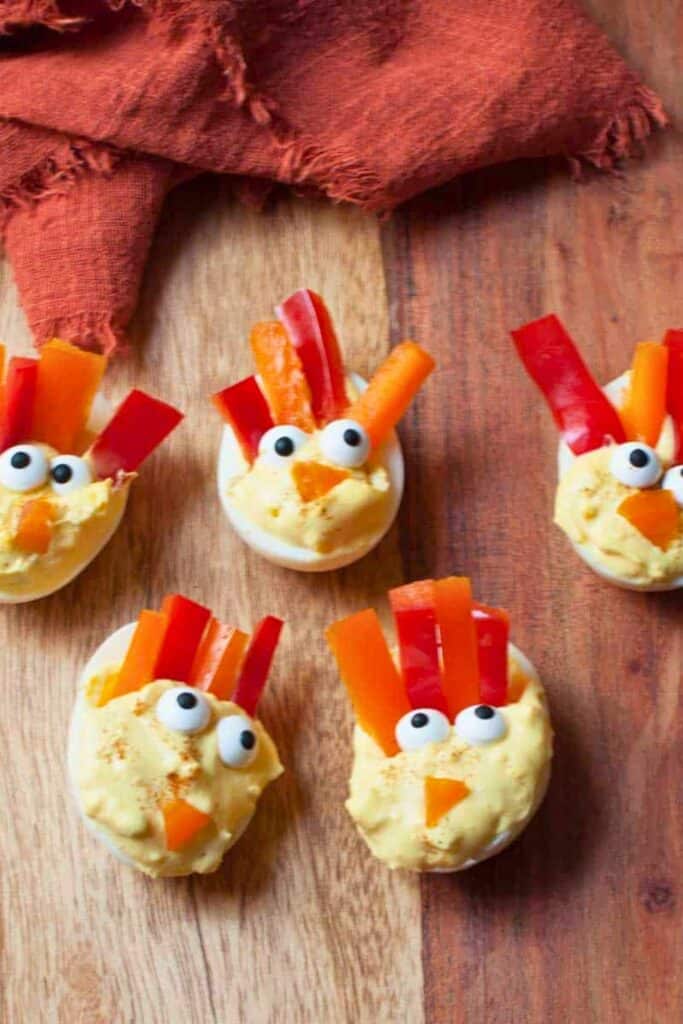 Turkey deviled eggs are arranged on a wooden board with creamy yellow filling decorated to resemble turkey faces. Candy eyes and orange pepper beaks create the faces while strips of red and orange bell pepper fan out behind each egg like colorful tail feathers.