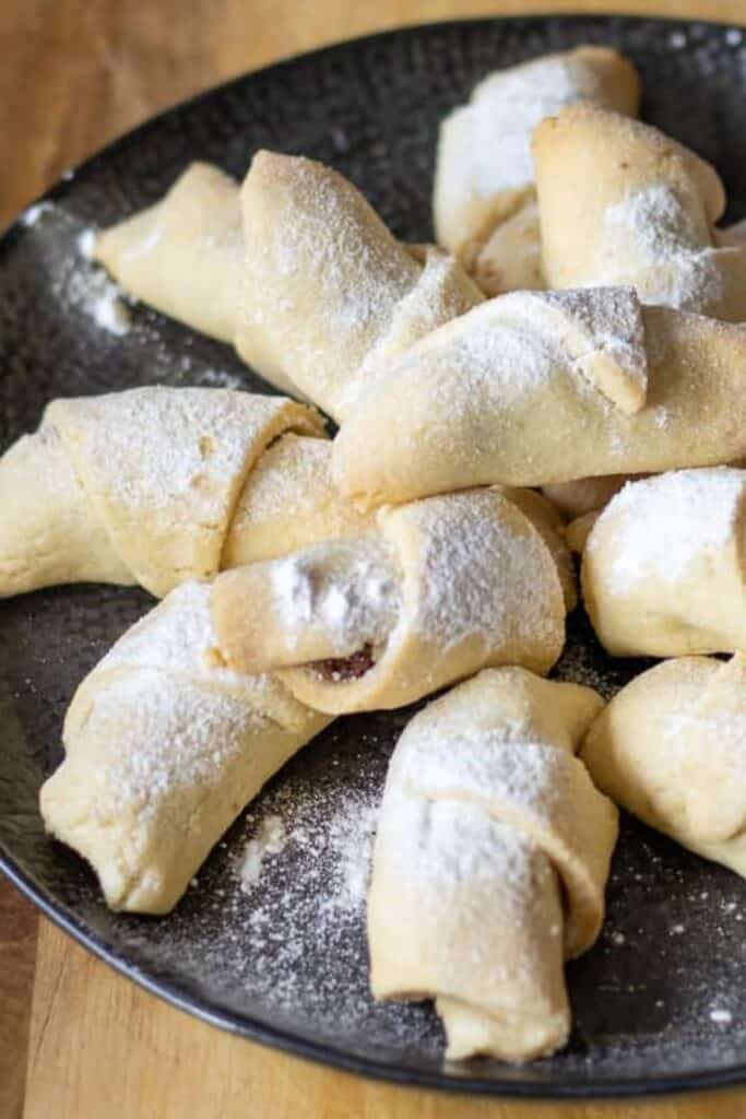 Turkish apple cookies dusted with powdered sugar and arranged on a dark plate. The soft pastry cookies are rolled around a sweet apple filling, with some pieces revealing the fruit center.