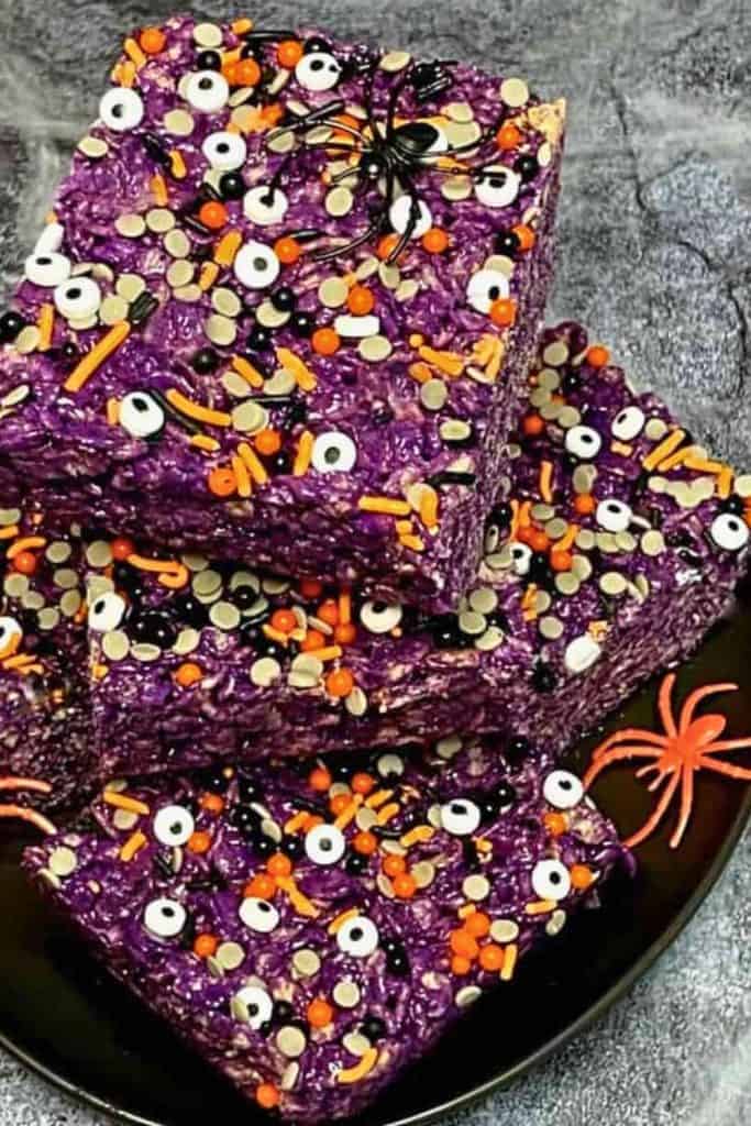 Purple Ube Rice Krispies treats topped with orange, black, and white Halloween sprinkles and candy eyes, garnished with plastic spider decorations on a dark plate.