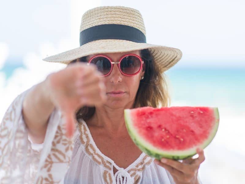 Middle aged woman with an angry face on the beach giving a thimbs down sign, holding watermelon.