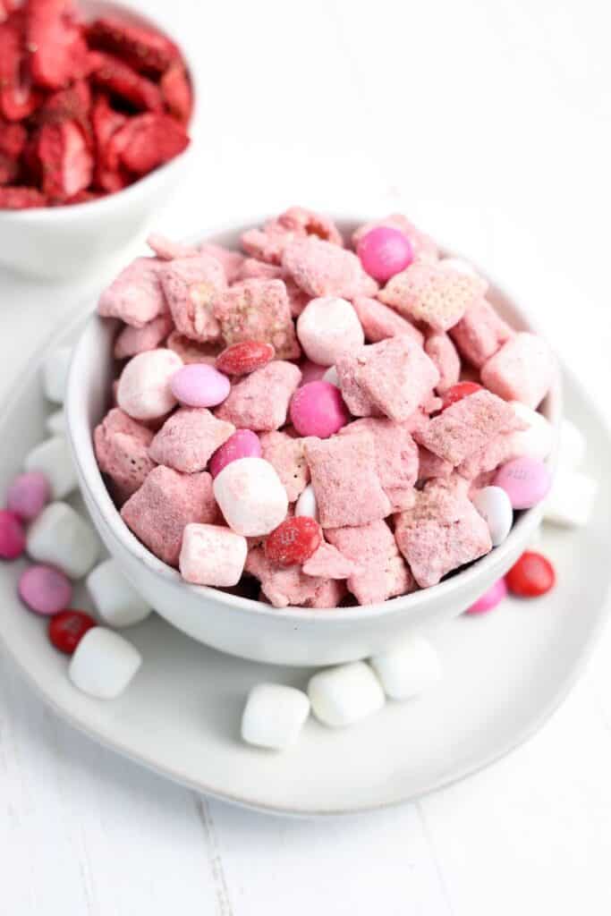 A close up of a medium sized bowl of Strawberry Puppy Chow snack mix with mini marshmallows and red, white and pink M&Ms with a bbowl of dried strawberries in the corner. 