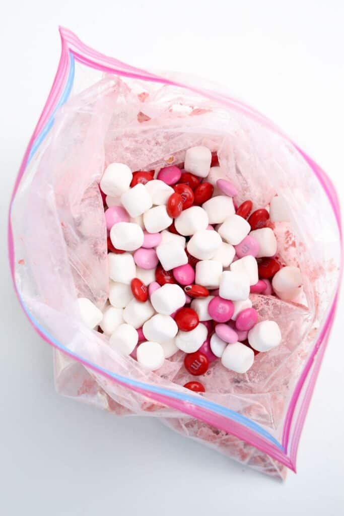 Overhead view of an open ziploc bag filled with strawberry, white chocolate covered Chex cereal, with mini marshmallows & M&Ms on top. 